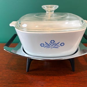 May include: A vintage white casserole dish with a clear glass lid, featuring a blue floral design. The dish sits on a chrome and black metal warming stand. A classic kitchenware item.