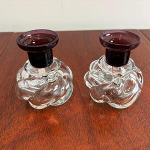 May include: Two clear glass perfume bottles with a knotted design. Each bottle has a dark purple stopper and a black neck. The bottles are displayed on a wooden surface.