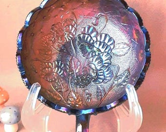Puede incluir: Un cuenco redondo de vidrio de carnaval azul oscuro iridiscente con un patrón floral moldeado de hojas y flores. El cuenco se exhibe sobre un soporte de plástico transparente, acompañado de tres pequeñas figuras decorativas de setas en rojo y blanco.