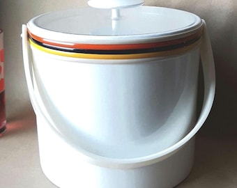 Puede incluir: Una cubitera cilíndrica blanca con tapa a juego y asa pivotante. El borde superior presenta rayas horizontales en naranja, negro y amarillo. Un vaso transparente con lunares rojos está situado a la izquierda de la cubitera.