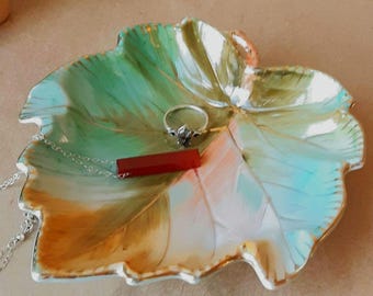 Puede incluir: Un plato de cerámica en forma de hoja con una mezcla de esmaltes verdes, blancos y marrones con bordes dorados. Un anillo de plata y un collar de plata con un colgante rectangular rojo descansan en el plato, junto a dos pequeñas figuras de setas.