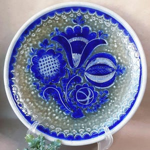 May include: A round ceramic plate with a blue floral design. The plate features a raised, detailed pattern of flowers and leaves in a deep blue color against a speckled gray background. The rim is white with a blue scalloped edge.
