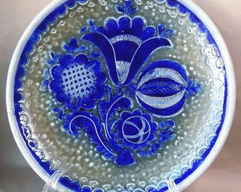 May include: A round ceramic plate with a blue floral design. The plate features a raised, detailed pattern of flowers and leaves in a deep blue colour against a speckled grey background. The rim is white with a blue scalloped edge.
