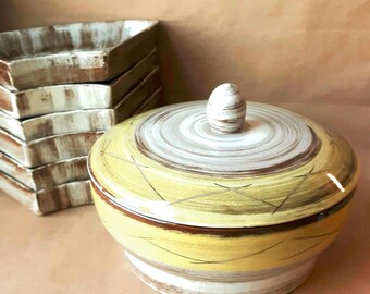 Puede incluir: Un cuenco redondo de cerámica con tapa, decorado con patrones de pinceladas horizontales en amarillo, marrón y blanco y un pequeño pomo, acompañado por una pila de seis platos triangulares de cerámica a juego con pinceladas marrones y blancas.