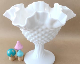 Puede incluir: Un cuenco de pedestal de vidrio blanco con un borde con volantes. Tres pequeñas figuras de hongos de madera de colores están frente al cuenco. Los hongos tienen tapas de color turquesa, rosa y azul.