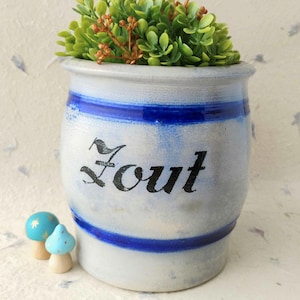 May include: A light gray ceramic crock with two horizontal cobalt blue bands and the word Zout written in black script. The container is filled with a cluster of green artificial foliage and is accompanied by two small light blue mushroom figurines.
