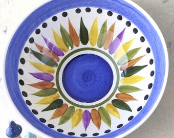 Puede incluir: Un cuenco de cerámica pintado a mano con borde y centro azules. El cuenco presenta un diseño de sol colorido con formas de hojas verdes, amarillas, moradas y marrones. Pequeños puntos negros rodean el sol. Dos pequeñas figuras de setas están junto al cuenco.