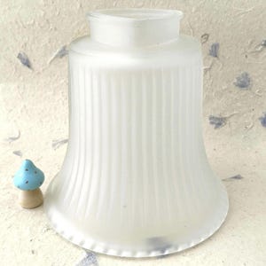 May include: A bell-shaped, frosted glass light shade featuring vertical ribbed detailing and a smooth, circular opening at the top. Beside the shade sits a small, light blue wooden mushroom figurine with a natural wood-toned base.