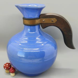 May include: A bright blue ceramic pitcher with a rounded body and a flared rim. The pitcher features a dark brown wooden handle attached to the neck with a brass-colored metal band secured by two screws. Two small red and white spotted mushrooms.