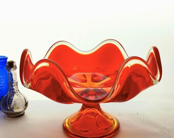 Puede incluir: Un cuenco de cristal naranja vibrante con un borde ancho, ondulado y festoneado, y una base de pedestal. Junto al cuenco hay una pequeña botella de cristal transparente con tapón de corcho y una botella alta de cristal azul cobalto con texto en relieve.