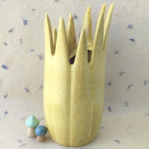 May include: A tall, yellow ceramic vase with a speckled finish and a jagged, crown-like top edge. Beside the base of the vase are two small, rounded mushroom-shaped ornaments, one light green with black dots and one blue with gold star accents.