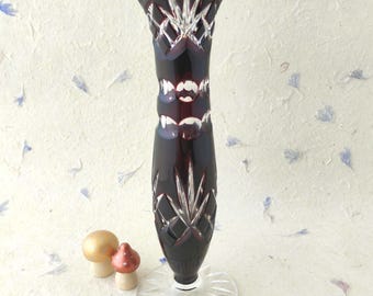 May include: A tall, slender, deep burgundy glass vase featuring intricate cut-glass patterns, including starburst motifs and circular indentations. The vase stands on a clear, circular, rayed glass base, showcasing a polished, reflective finish.