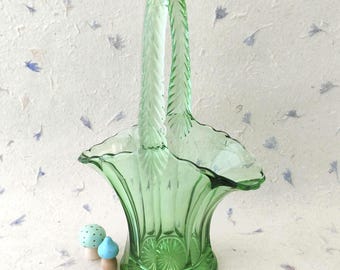 May include: A light green glass basket featuring a tall, textured handle with a feather-like pattern, a scalloped rim, and a vertical ribbed body that flares outward. The base displays a sunburst design, and two small mushroom figurines sit nearby.