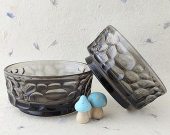 May include: Two round, dark grey glass bowls featuring a textured, hammered surface pattern. The bowls are positioned with one sitting upright and the other tilted on its side. Two small, light blue wooden mushroom figurines sit in front.