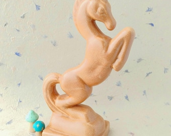 Puede incluir: Una figura de caballo de cerámica vidriada de color tostado en pose encabritada, montada sobre una base rectangular. Junto al caballo hay dos pequeños adornos redondeados en forma de seta, uno verde claro y otro azul, con un pequeño patrón de estrella dorada.
