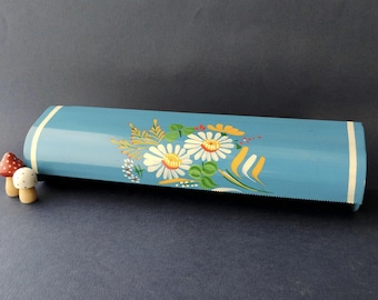 May include: A light blue metal kitchen foil or plastic wrap dispenser featuring a painted floral design of white daisies with yellow centres and green leaves, accented by thin white vertical stripes on the left and right edges of the unit.