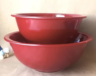 May include: Two red ceramic mixing bowls, one stacked on top of the other. The bowls have a glossy finish and a rounded shape. The bowls are likely for kitchen use.