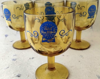 Puede incluir: Un juego de tres copas de cristal de color ámbar, cada una con un logotipo azul impreso que dice Pabst Blue Ribbon Beer con un gráfico de cinta y remolinos decorativos. Las copas tienen un cuenco redondeado, un tallo corto y una base circular.