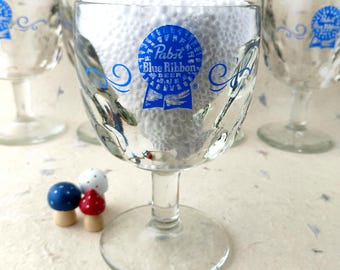 Puede incluir: Una copa de cristal transparente con el logotipo azul de Pabst Blue Ribbon Beer y adornos de volutas. El cristal tiene una superficie texturizada con hoyuelos, un tallo corto y una base circular. Pequeños adornos en forma de seta están cerca.