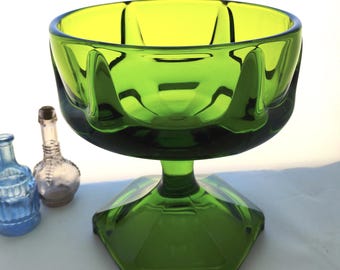 May include: A green glass pedestal bowl with a hexagonal base and a scalloped rim. The bowl is a vibrant green colour and has a clear, glossy finish. Two small decorative glass bottles are in the background.
