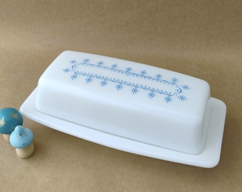 May include: A white rectangular butter dish with a lid featuring a blue snowflake and scalloped line pattern. The dish has a matching white base with a flat rim. Two small blue mushroom-shaped decorative items sit to the left of the dish.