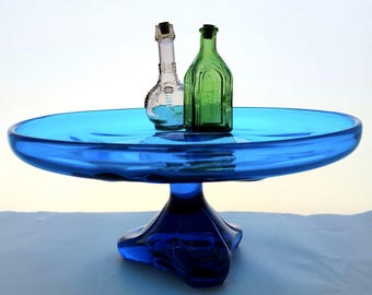 May include: A striking blue glass cake stand with a broad, shallow dish and a pedestal base. Two small glass bottles with cork stoppers are on the stand. One bottle is clear, the other is green.