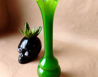 May include: A striking green glass vase with a flared, ruffled top. The vase has a rounded base and a long, elegant neck. A black skull-shaped planter with a succulent is next to it.