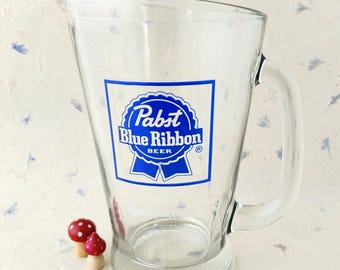 Puede incluir: Una jarra de cerveza de cristal transparente con el logotipo azul de Pabst Blue Ribbon impreso en el frente. La jarra tiene un asa resistente, un pico vertedor ancho y una base gruesa. El texto Quality Since 1844 aparece en letras blancas.