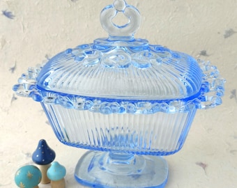 Puede incluir: Un dulcero de vidrio prensado azul claro con forma rectangular, acanalado vertical y una tapa de borde festoneado con un asa central. Tres pequeñas figuras decorativas de setas de madera en azul y blanco se encuentran al lado.