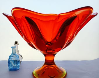 May include: A striking orange glass bowl with a pedestal base. The bowl has a distinctive, petal-like design. Two small glass bottles are in the background, one blue and one clear. The bowl is a vibrant shade of orange.