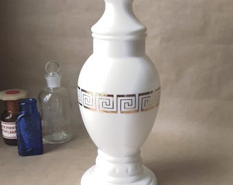 Puede incluir: Un frasco de boticario de vidrio lechoso blanco con cuello estrecho y base ancha. El centro del frasco presenta una banda horizontal con un patrón de llave griega en oro metálico. El frasco tiene un acabado liso y brillante y una forma bulbosa.