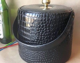 May include: Black ice bucket with a textured, crocodile-like pattern. It has a gold-coloured knob on the lid and a black handle. The bucket is cylindrical and appears to be made of a durable material. A green bottle is partially visible.