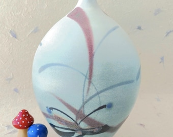 May include: A pale blue ceramic vase with abstract floral patterns in pink, blue, and black. Two small wooden mushroom figurines, one red with white spots and the other blue with white spots, are in front of the vase.