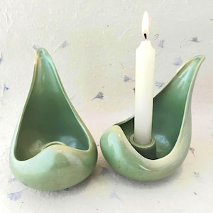 Puede incluir: Dos candelabros de cerámica verde con diseño de lágrima. Un portavelas contiene una vela blanca encendida. Los candelabros tienen un acabado liso y brillante y una forma orgánica única. El fondo es una superficie clara y texturizada.