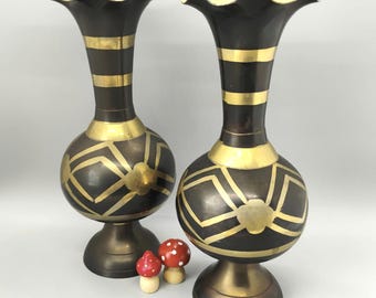 May include: Two decorative black and gold vases with a unique shape and design. The vases have a flared top with a gold rim, gold bands, and geometric patterns. Two small wooden mushroom figurines are in front of the vases.