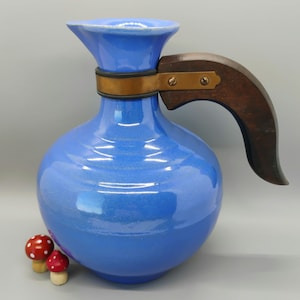 May include: A blue ceramic pitcher with a wooden handle and a brass band around the neck. The pitcher has a rounded body and a flared spout.