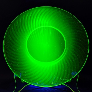 May include: A glowing green glass plate with a spiral design. The plate is illuminated, showcasing its texture and shape. The plate is on a clear stand against a black background.