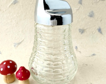 Puede incluir: Un azucarero de cristal transparente con un patrón texturizado de costillas horizontales y una tapa de metal cromado pulido, acompañado por dos pequeños adornos de setas de madera con capuchón rojo y puntos blancos.
