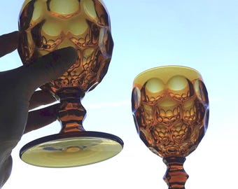 May include: Two amber-coloured glass goblets featuring a prominent honeycomb or dimpled texture across the bowl. Each piece has a short, faceted stem and a wide, circular base, crafted from translucent glass that catches the light.