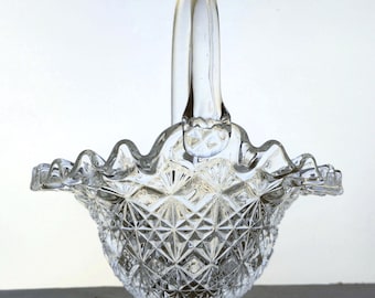 May include: A clear glass basket featuring a diamond-cut pattern on the exterior body, a ruffled scalloped rim, and a single arched handle. The base is circular and pedestal-style, supporting the textured bowl of the decorative glass vessel.