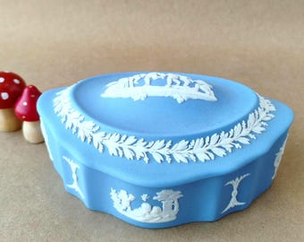 May include: A light blue oval trinket box featuring white relief ornamentation. The lid displays a central scene of figures, while the sides are decorated with a continuous leafy garland and small vignettes of cherubs and urns in white relief.