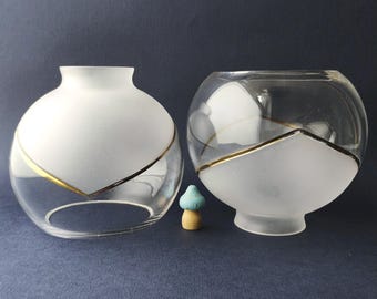 May include: Two spherical glass lamp shades featuring a combination of clear and frosted glass sections separated by a thin gold-coloured line. A small, light blue and tan mushroom-shaped figurine is positioned between the two glass shades.