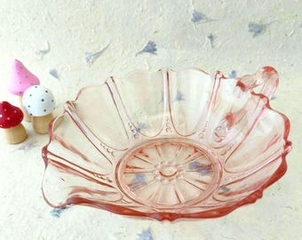 Puede incluir: Un cuenco de cristal rosa translúcido con borde festoneado y un patrón radial prensado en la base. El cuenco presenta una pequeña asa en un lado y detalles decorativos en forma de cuentas a lo largo de las nervaduras verticales interiores.