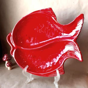 May include: A red ceramic fish-shaped serving dish with two compartments. The dish is glazed and has a glossy finish. It is displayed on a clear plastic stand.