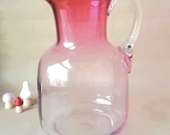 Puede incluir: Una jarra de vidrio transparente con un efecto degradado rosa, con un asa curva y un pico vertedor. La jarra está acompañada de pequeñas figuras decorativas de setas. Es un artículo decorativo.