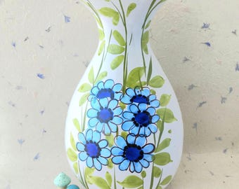Puede incluir: Un jarrón de cerámica blanca con un diseño pintado a mano de flores de color azul brillante con centros azul oscuro y hojas de color verde claro sobre tallos largos. El jarrón tiene un borde ancho y acampanado y una base cónica, junto a dos pequeñas figuras de setas azules.