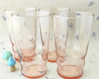 May include: A set of four tall, light pink drinking glasses with a textured, wavy surface. The glasses are clear and have a cylindrical shape with a slightly flared rim. Two small, decorative mushroom-shaped ornaments are placed nearby.