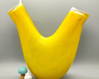 Puede incluir: Un jarrón de vidrio amarillo brillante con un diseño curvo único. El jarrón tiene un acabado liso y brillante y parece estar hecho a mano.
