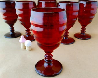 May include: A set of six deep red glass goblets featuring a horizontal ribbed texture on the bowl, a short stem with a decorative ringed detail, and a circular base. Two small, light-coloured mushroom-shaped ornaments sit near the front glass.