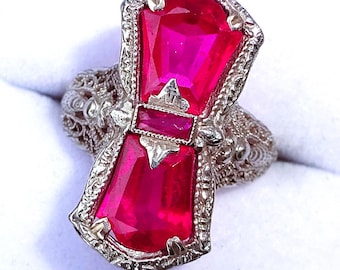 May include: A silver filigree ring with a large, faceted, ruby-red gemstone set in the center. The ring has a delicate, intricate design.